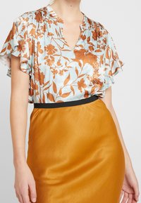 Kvinde iført en lyseblå bluse med brun blomsterprint og flæseærmer, stoppet ned i en højtaljet, sennepsgul nederdel med sort talje.