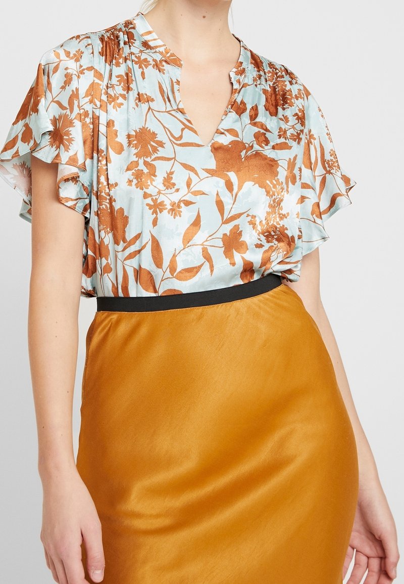 Kvinde iført en lyseblå bluse med brun blomsterprint og flæseærmer, stoppet ned i en højtaljet, sennepsgul nederdel med sort talje.