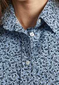 Camisa de color azul claro con un patrón floral en negro y acentos en blanco, cuello tipo botón y dos botones visibles en la parte delantera.