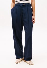 Marinblå veckade byxor med skräddarsydd passform, utrustade med sidofickor och en knappstängning. Bärs med beige vävda slip-on-skor.