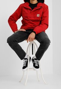 Red lightweight jacket with hood, front pocket, and Carhartt logo. Paired with black jeans and black-and-white sneakers, seated on a white stool.
