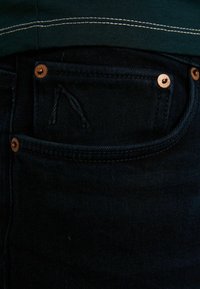 Jeans de mezclilla oscura que presentan un bolsillo frontal con remaches de color cobre y un detalle de logo bordado, combinados con una camisa verde oscuro.