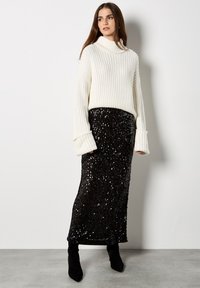 Fleeciger Creme-Turtleneck-Pullover mit geribbelter Textur, kombiniert mit einem langen schwarzen Paillettenrock. Schwarze Stiefeletten runden das Outfit ab.