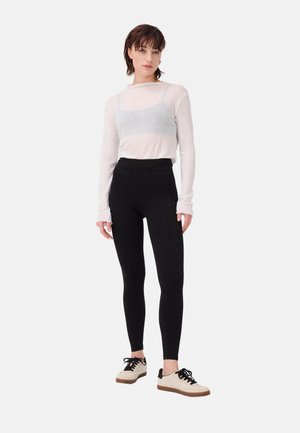 Vrouw staat en draagt een doorschijnende witte top met lange mouwen, een grijze bralette, zwarte hoge taille leggings en witte sneakers met zwarte veters.