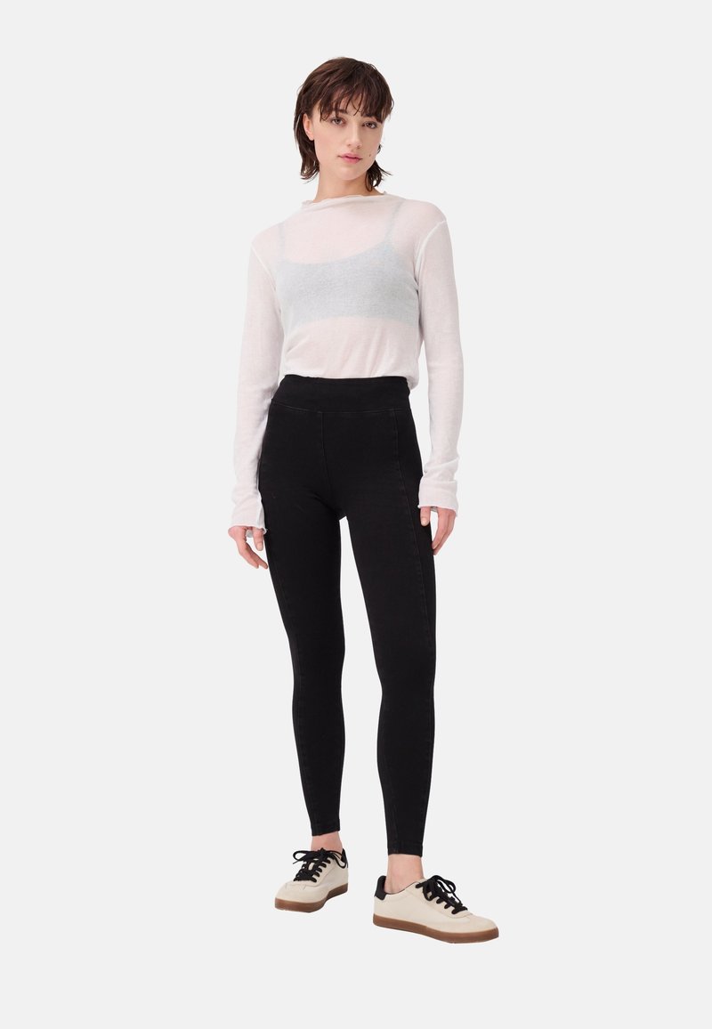 Woman standing wearing a sheer white long-sleeve top with a gray bralette, black high-waisted leggings, and white sneakers with black laces.