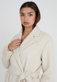 Cappotto avvolgente texture color crema con colletto a rever e cintura, dotato di maniche lunghe e materiale morbido e peloso.