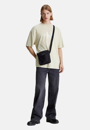 Bolso cruzado negro con un acabado texturizado y parche con el logotipo. Lleva puesto una camiseta verde claro y vaqueros oscuros holgados. Botas negras de suela gruesa.