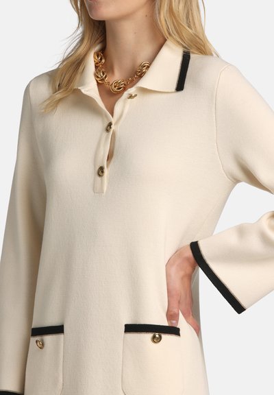 Béžový pletený top s čiernym lemovaním na golieri, manžetách a vreckách, zlatými gombíkmi a hrubým zlatým reťazovým náhrdelníkom na žene s jednou rukou v bok.