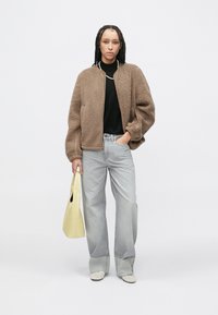 Bruine fleece bomberjack met een ritssluiting aan de voorkant, gecombineerd met een zwart overhemd en lichtgrijze jeans. Lichtgele draagtas en beige schoenen maken de look compleet.