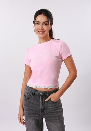 Νέα γυναίκα με σκούρα μαλλιά σε αλογοουρά, φοράει ένα ανοιχτό ροζ κοντό μπλουζάκι με δαντέλα και σκούρα γκρι τζιν, χαμογελάει με το χέρι στην τσέπη.