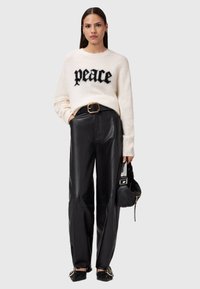 Biały dziany sweter z napisem „peace” w czerni, zestawiony z czarnymi skórzanymi spodniami z wysokim stanem. Modelka trzyma małą czarną torebkę.