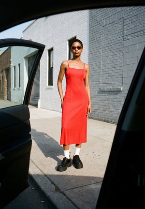 Frau trägt ein rotes Kleid, Sonnenbrille, weiße Socken und schwarze Plateauschuhe und steht auf dem Gehweg neben einem grauen Ziegelgebäude.