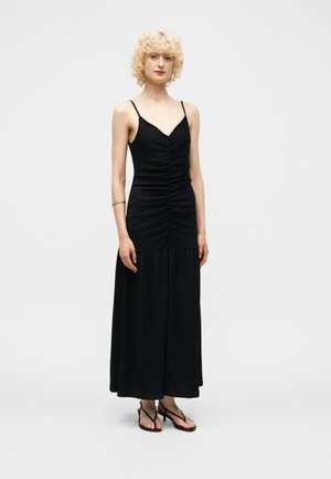 Γυναίκα με ξανθά σγουρά μαλλιά, που φοράει μακρύ μαύρο φόρεμα με λεπτές τιράντες και μαύρες ανοιχτές γόβες σε μονόχρωμο φόντο.