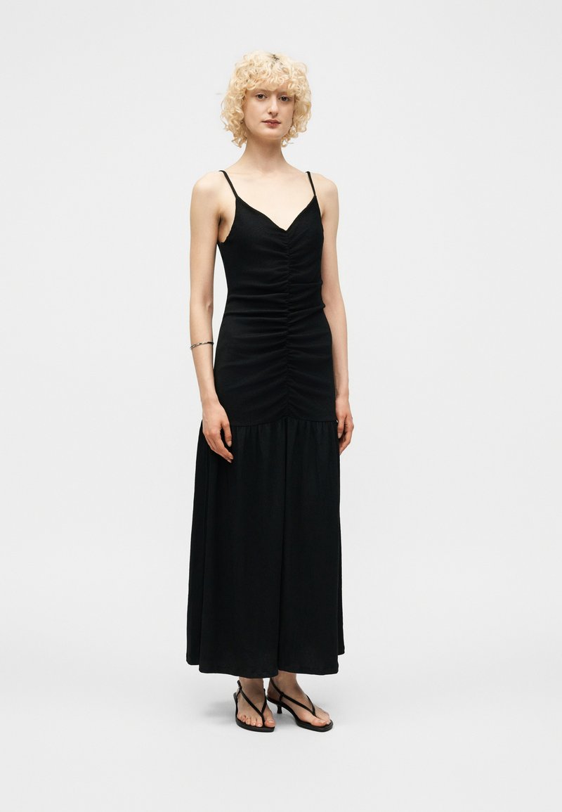 Γυναίκα με ξανθά σγουρά μαλλιά, που φοράει μακρύ μαύρο φόρεμα με λεπτές τιράντες και μαύρες ανοιχτές γόβες σε μονόχρωμο φόντο.