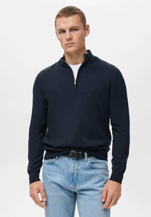 Marineblauwe trui met ritssluiting in een zachte stof, voorzien van lange mouwen en een relaxed fit, gecombineerd met lichtblauwe jeans en een zwarte riem.