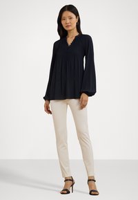 Marineblauwe blouse met pofmouwen, geplooid detail en een V-hals, gecombineerd met beige aansluitende broeken en zwarte schoenen met stilettohak.