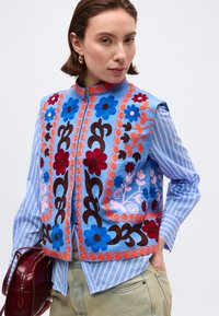 Γυναίκα που φοράει μπλε γιλέκο κεντημένο με λουλούδια πάνω από μία ριγέ πουκάμισο, κρατώντας μία μπορντό τσάντα, ποζάροντας μπροστά σε απλό φόντο.