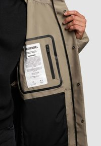 Interior de chaqueta caqui con un bolsillo negro, cremallera y una etiqueta con texto. Tejido suave y detalle en contraste.