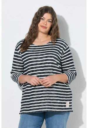 Junge Frau mit welligem braunem Haar, die ein schwarz-weiß gestreiftes Langarmshirt und blaue Jeans trägt, steht an einer weißen Wand.