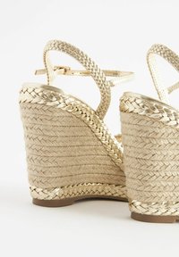 Sandales compensées beiges tissées avec garniture tressée dorée et brides à la cheville sur fond blanc.
