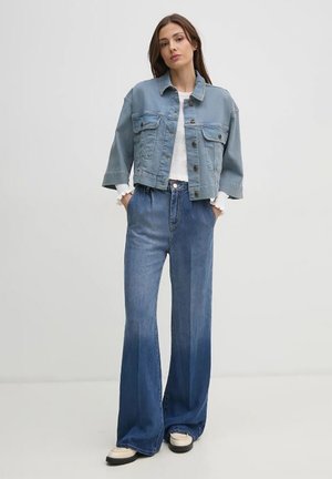 Giacca di jeans blu chiaro adatta, con tasche a toppa, abbinata a jeans blu a vita alta svasati. La modella indossa sneaker crema.