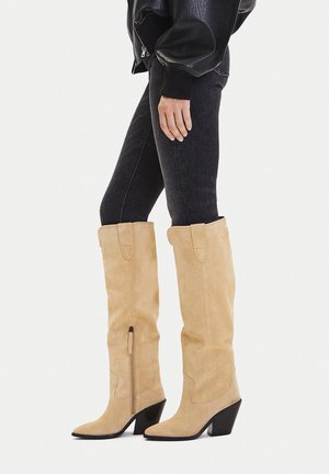 Person wearing beige suede knee-high heeled boots with side zippers, dark skinny jeans, and a black leather jacket.