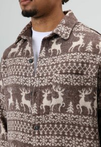 Chemise en flanelle marron avec motifs de cerfs blancs et flocons de neige. Comprend un col, une fermeture à boutons et une poche poitrine.