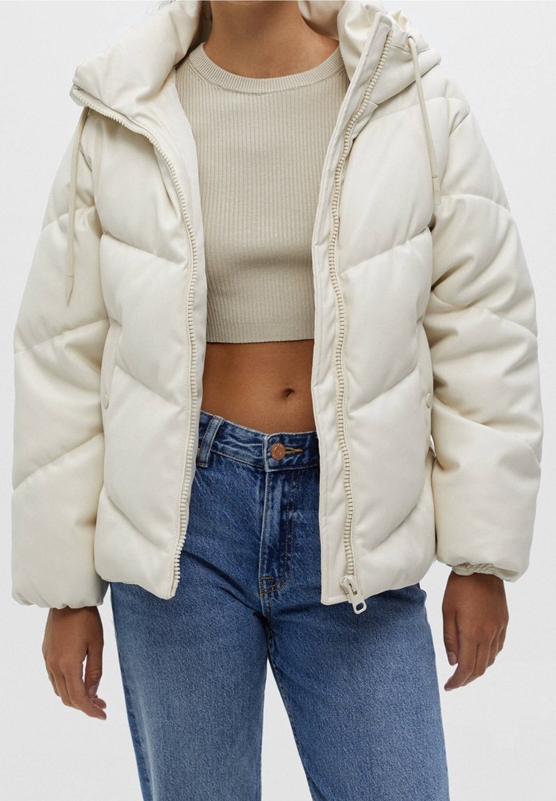 Personne portant une doudoune crème par-dessus un top court côtelé beige et un jean bleu taille haute sur un fond uni.