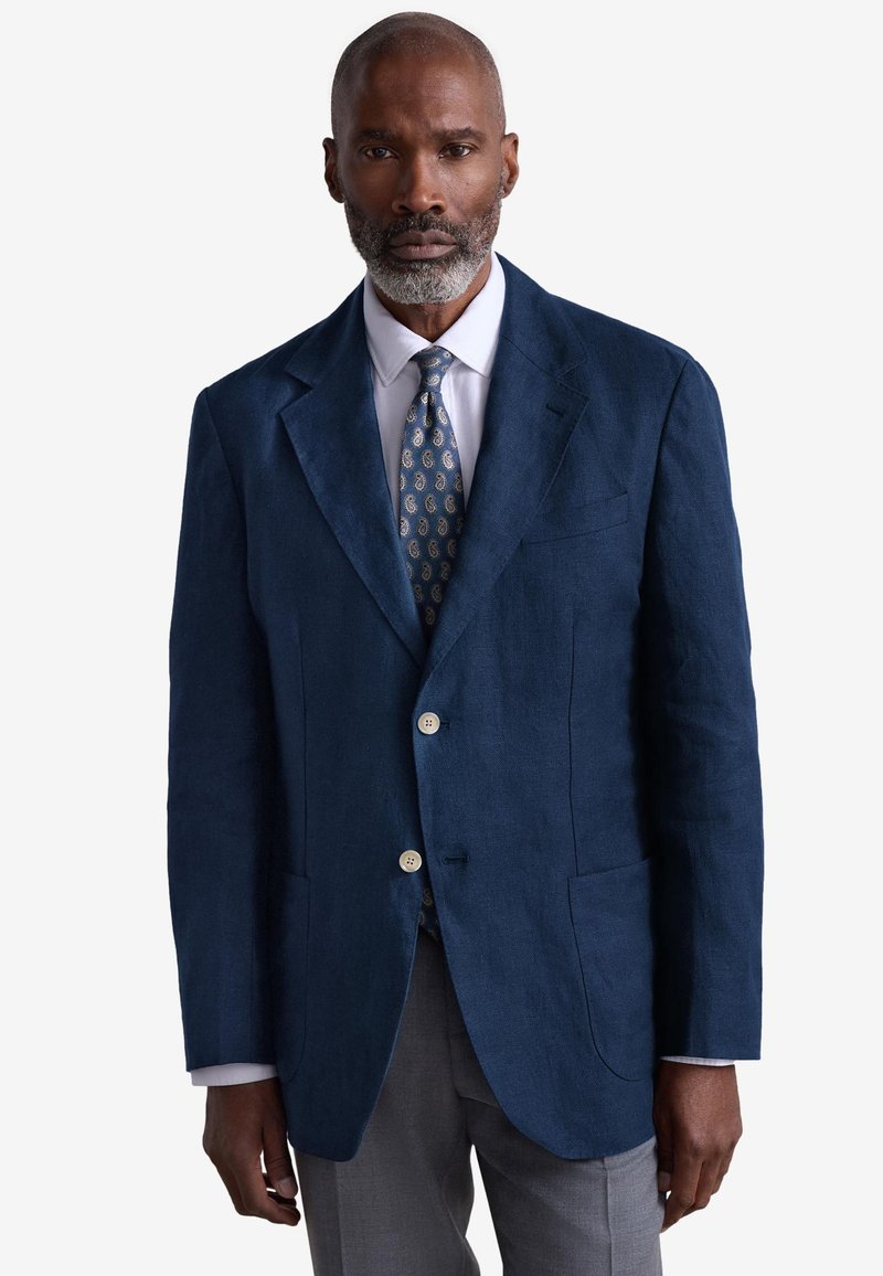 Marineblauer Leinenblazer mit zwei Vordertaschen, zwei Knöpfen und Notch-Revers. Weißes Hemd und gemustertem Krawatte runden den Look ab.