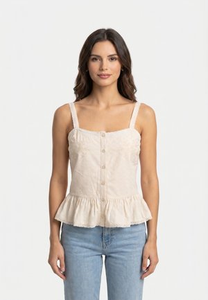 Femme aux cheveux bruns ondulés porte un haut beige sans manches boutonné avec un ourlet à volants et un jean bleu clair, debout sur fond blanc.