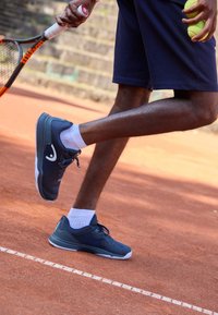 Ναυτικό μπλε αθλητικά παπούτσια με λευκές λεπτομέρειες, με υφή στην επιφάνεια και υποστηρικτικό σχέδιο, συνδυασμένα με λευκές κάλτσες και μαύρες βερμούδες σε χωμάτινη επιφάνεια.