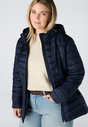 Femme blonde portant une veste matelassée bleu marine avec capuche sur un pull beige et un jean bleu clair, une main dans la poche.