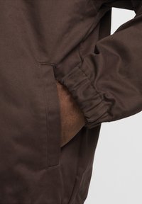Brun jakke stof med en glat tekstur; har et elastisk manchett og sidelomme. Designet fremhæver funktionalitet og komfort.