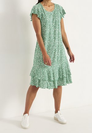 Femme portant une robe verte à fleurs longueur genou avec manches courtes volantées et ourlet à étages, associée à des baskets blanches, debout contre un mur uni.