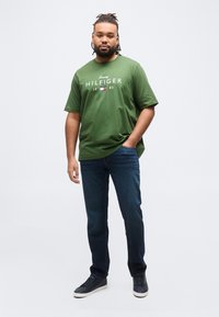 Άνδρας με κοτσίδες, φοράει πράσινο μπλουζάκι Tommy Hilfiger, σκούρο τζιν και μαύρα αθλητικά παπούτσια, στέκεται με ένα χέρι στην τσέπη μπροστά σε λευκό φόντο.