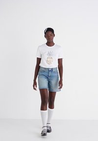 T-shirt bianco con grafica di ananas, abbinato a pantaloni shorts in denim azzurro. Il modello indossa calze bianche al ginocchio e sandali neri. Sfondo semplice.