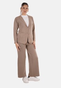 Completo beige a due pezzi con blazer su misura a chiusura con un solo bottone, abbinato a pantaloni a gamba larga. Il modello indossa sneakers bianche.