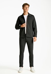 Chaqueta negra de botones con un bolsillo en el pecho, combinada con pantalones negros ajustados. El modelo lleva una camiseta blanca y zapatillas blancas. Diseño minimalista.