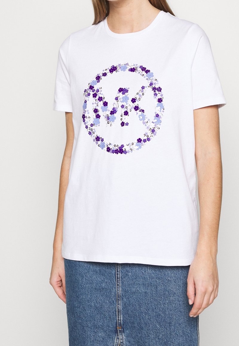 T-shirt en coton blanc arborant un motif floral dans des nuances de bleu et de violet, avec un logo circulaire au centre. Manches courtes, coupe décontractée.