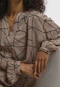 Blouse beige à motifs avec un design géométrique noir, un col en V, une patte de boutonnage et des poignets de manches froncés.