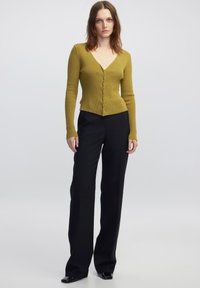 Cardigan ribbed verde oliva con scollo a V e chiusura a bottoni, abbinato a pantaloni neri a vita alta e scarpe nere eleganti.