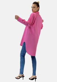 Langärmliges, leuchtend pinkes Shirt mit lockerer Passform und abgerundetem Saum, kombiniert mit blauen Skinny-Jeans und schwarzen High-Heelsandalen.