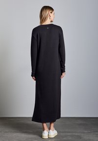 Svart, långärmad maxiklänning med rund hals och mjuk textur. Har en liten logotyp nära halsringningen. Bärs med vita sneakers.