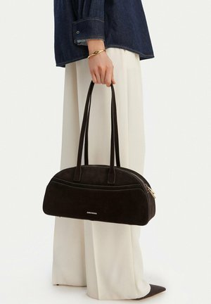 Mano sosteniendo un bolso negro de Gino Rossi con correas largas, con pantalones blancos, chaqueta oscura, pulsera dorada y zapatos negros puntiagudos.