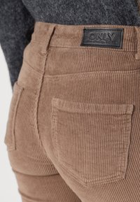 Bruna kordbyxor har vertikala ränder, en svart etikett med "ONLY" på midjebandet och två bakfickor. En grå tröja är delvis synlig.