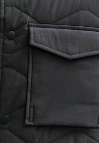 Czarna pikowana kurtka z tkaniny, z naszywaną kieszenią, zapięciem na zatrzask oraz gładką fakturą z trójkątnym wzorem przeszycia.