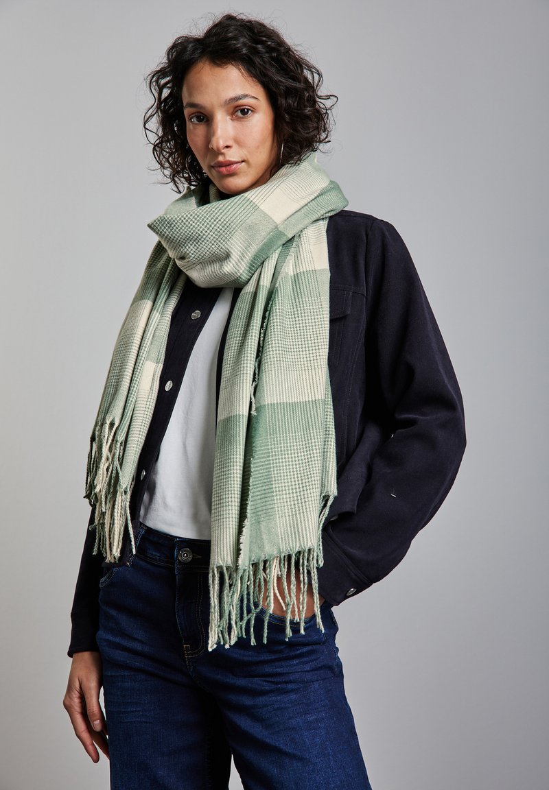 Scarf a scacchi verde e crema realizzata in morbido tessuto, con bordi frangiati. Indossato sopra una giacca scura e una camicia bianca.