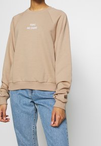 Sweat-shirt en coton beige avec col rond et manches raglan. Texte centré : « PEOPLE ARE STRANGE. » Ourlet et poignets côtelés pour un meilleur ajustement.