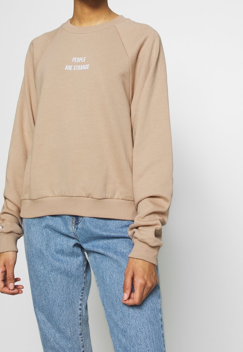 Sweat-shirt en coton beige avec col rond et manches raglan. Texte centré : « PEOPLE ARE STRANGE. » Ourlet et poignets côtelés pour un meilleur ajustement.