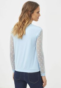 Suéter azul claro con un cuerpo liso y mangas de malla transparente que presentan lunares azules claros. Puños y dobladillo acanalados completan el diseño.